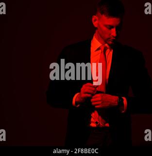 Sad thoughtful man in official suit stands looking down at corner over black background. Red light filter, gel Stock Photo
