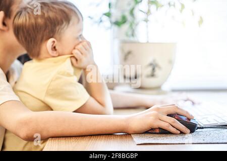 Concentrated little boy in yellow t-shirt looks into computer sitting on brother knees holding hands on mouse and keyboard at home Stock Photo