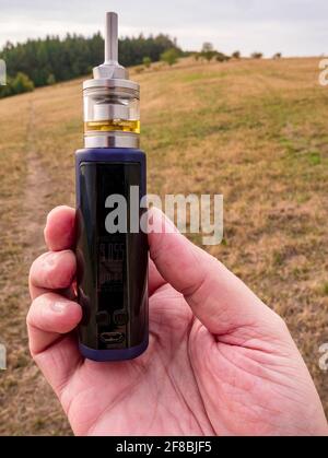Hand holding vaping mod with tank filled with liquid. In background footpath on meadow and hill with forest. Stock Photo