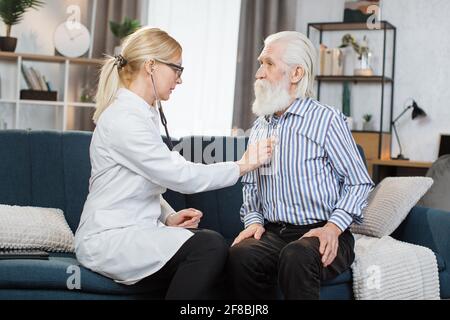 Senior calm bearded man sitting on the couch while caring experienced nice female doctor listening his heartbeat during home visit,front view Stock Photo