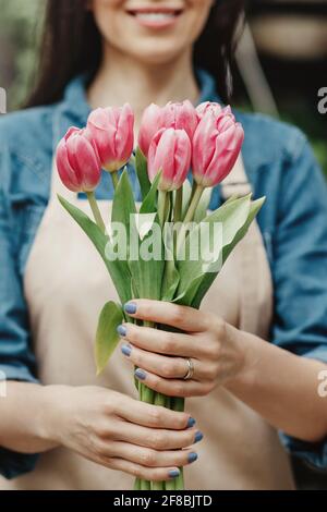 Cheerful charming young woman florist, owner standing and holding pink tulips in shop, recommends flowers Stock Photo