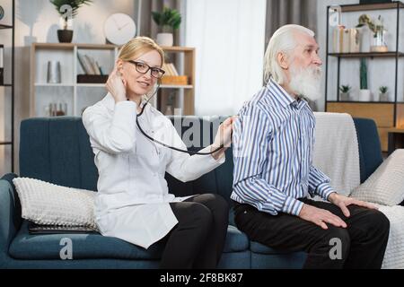 Female physician listening patient back through stethoscope, examining heart beat and lungs of senior bearded man during home visit. Geriatric heart and lungs deseases. Stock Photo