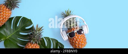 Funny pineapple wearing white headphone, concept of listening music, isolated on blue background with tropical palm leaves, top view, flat lay design. Stock Photo