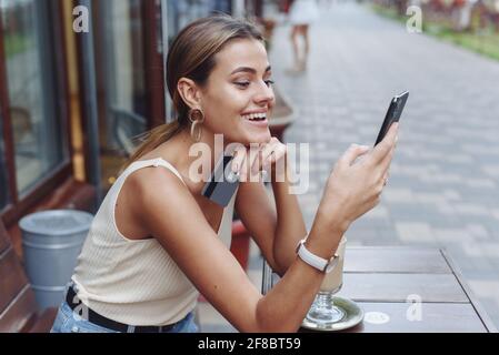 Young smiling woman shopping online using phone outdoors in cafe. A beautiful model looks at the phone and drinks coffee in the summer. Stock Photo