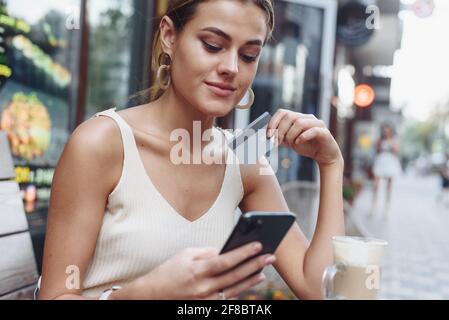 Woman sitting at cafe holding mobile phone and plastic card signing up on website.Beautiful model paying with credit card while shopping online using Stock Photo