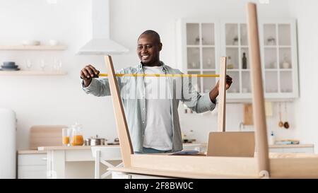 Excited black man measuring furniture with roulette, assembling new table in kitchen interior Stock Photo