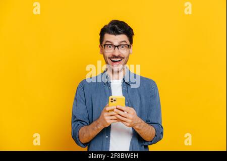 Happy handsome amazed stylish guy with eyeglasses, using smartphone, chatting online, writing message, browsing internet, social media, looks surprised at camera, standing on isolated orange Stock Photo