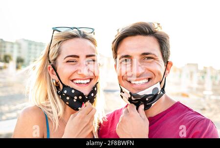 Young couple in love smiling with open face mask - New normal life style and relationship concept with happy lovers on positive mood at beach Stock Photo