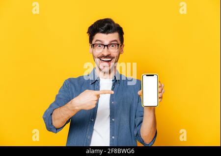 Happy charismatic unshaven caucasian guy in denim shirt and glasses holds smartphone with blank white screen for your presentation and points finger at it, stands on isolated orange background, smiles Stock Photo