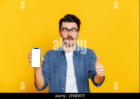 A funny stylishly dressed guy with foolish facial expression holds smartphone with blank white screen for your presentation, shows thumb up, stands on isolated orange background Stock Photo