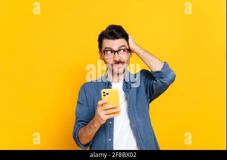 Confused puzzled caucasian unhaved guy, stylishly dressed, holding a smartphone in his hand, looking pensively to the side, holding his head with his hand, standing an isolated orange background Stock Photo