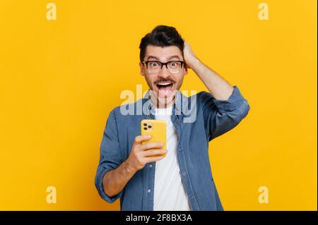 Amazed shocked positive caucasian guy with glasses, uses smartphone, browsing Internet, social networks, get unexpected news, holding his head with his hand, stands on isolated orange background Stock Photo