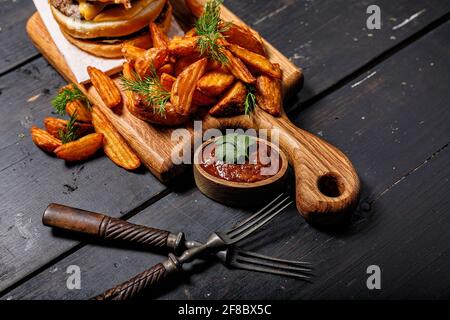 Appetizing fried potato wedges and burger on a wooden cutting board. Big portion of baked potatoes with herbs and tomato sauce. Fast food foodporn Stock Photo