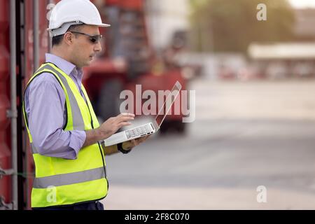 Confident Caucasian man engineer wearing white safety helmet using computer laptop and check for control loading containers box from Cargo freight shi Stock Photo