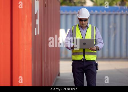 Confident Caucasian man engineer wearing white safety helmet using computer laptop and check for control loading containers box from Cargo freight shi Stock Photo