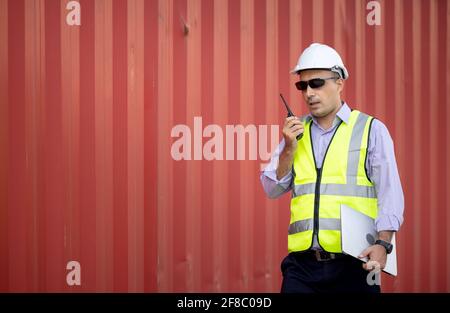 Man engineer using walkie-talkie in shipping yard, Industrial worker is controlling container loading by walkie talkie in import-export business. Stock Photo