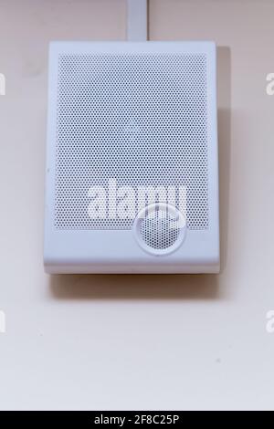 A white radio transmitter hangs on a beige wall. On the front side there is a grill with small holes for a loudspeaker. Electronic device for listening to messages, music and news. Stock Photo