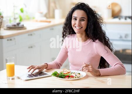 Portraif Of Happy Smiling Young Lady Having Breakfast And Using Digital Tablet Stock Photo