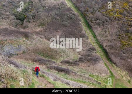A woman in red coat walks in the countryside down a steep path through lush green fields and hills Stock Photo