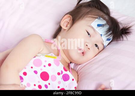 Close-up little asian girl lying  on sickbed with mercury thermometer in her armpit and cooling fever patch on her forehead Stock Photo