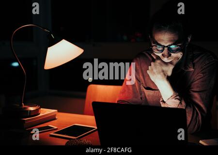 Tired caucasian male freelancer sorting through images on laptop working late at night in home office Stock Photo