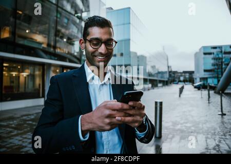 Mixed race businessman smiling while texting on smartphone walking on his way to work  Stock Photo