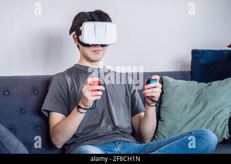 Gaming at home with a VR headset, Virtual Reality, teenager / young man in his twenties, living room setting, sitting on a sofa Stock Photo