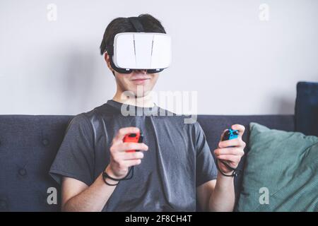 Gaming at home with a VR headset, Virtual Reality, teenager / young man in his twenties, living room setting, sitting on a sofa Stock Photo