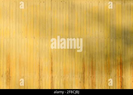 Wooden boards painted with yellow paint are fading to reveal an orange coasting below.  Nail holes are visible in three rows and a tree casts shadows Stock Photo