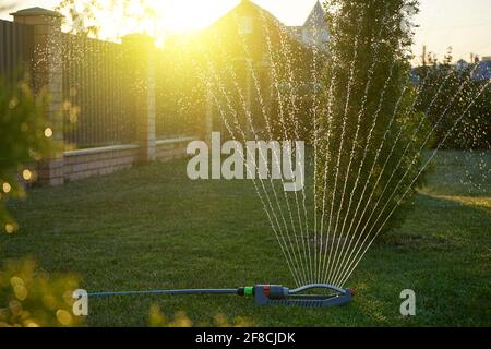 A sprayer waters the lawn in the evening against the backdrop of a summer sunset Stock Photo