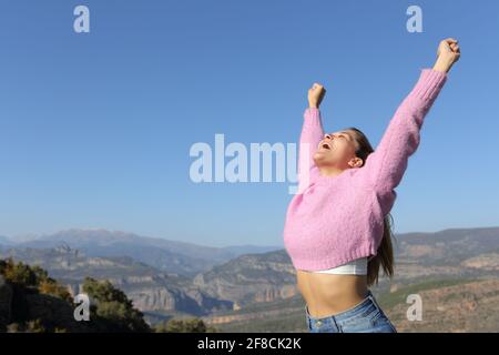 Excited woman in pink raising arms celebrating vacation in the mountain a sunny day Stock Photo