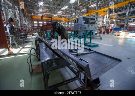 Locomotive-building plant workshop. Two Asian welder workers make metal part. Locomotive cabin and worker on background. Stock Photo
