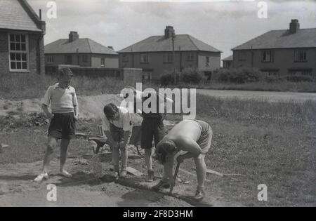 1963, historical, summertime and standing on a empty plot of land on a semi-rural housing estate, three curious young lads looking at a man digging out a drainage channel on a domestic building site, perhaps for a garage, England, UK. Stock Photo