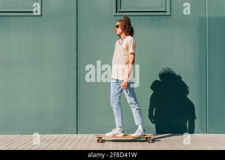 horizontal portrait of a young skateboarder riding his skate on a green background. He has long curly hair and wears sunglasses Stock Photo