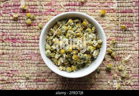 Dry chamomile flowers in a bowl on woven cotton rustic pink fabric, from above overhead top view, flat lay, closeup, naturopathy, homeopathy and natur Stock Photo