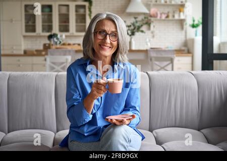 Senior smiling 60s woman portrait indoors looking at camera with cup of tea, coffee. Stock Photo