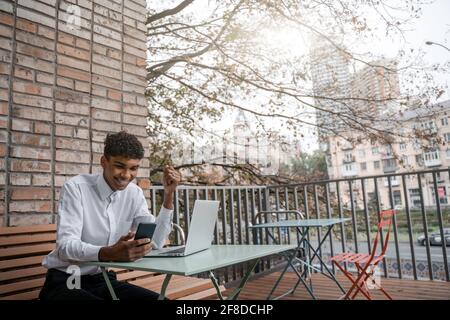 A black man sits on a summer terrace or veranda near a cafe. The guy works at the laptop. View of the spring business downtown Stock Photo