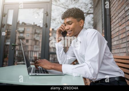 Cheerful man works at a laptop and speaks on the phone. A black guy sits on a summer terrace or veranda near a brick wall. View of the spring business Stock Photo