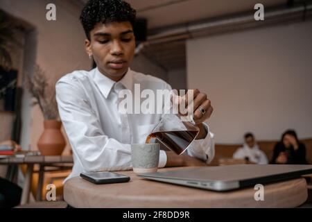 A young black man pours tea from a teapot into a cup. The guy in the cafe. Work outside the office, remote work or study concept. Stock Photo