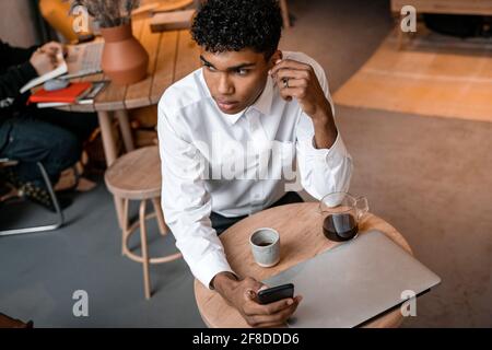 A young black man is sitting at the table with a laptop, drinking tea and using a smartphone. The guy in the cafe. Work outside the office, remote Stock Photo