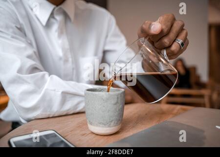 A young black man pours tea from a teapot into a cup. The guy in the cafe. Work outside the office, remote work or study concept. Stock Photo