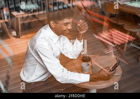 A young black man is sitting at the table with a laptop, drinking tea and using a smartphone. The guy in the cafe. Work outside the office, remote Stock Photo