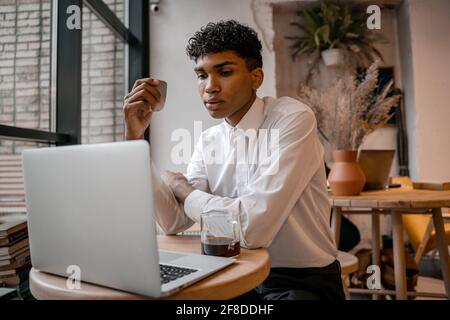 A young black man is sitting at the table with a laptop, drinking tea and using a smartphone. The guy in the cafe. Work outside the office, remote Stock Photo