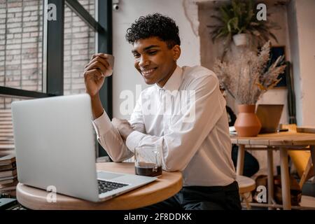 A young black man is sitting at the table with a laptop, drinking tea and using a smartphone. The guy in the cafe. Work outside the office, remote Stock Photo