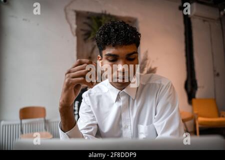 A young black man is sitting at the table with a laptop, drinking tea and using a smartphone. The guy in the cafe. Work outside the office, remote Stock Photo