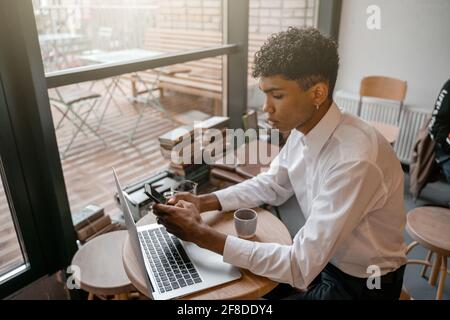 A young black man is sitting at the table with a laptop, drinking tea and using a smartphone. The guy in the cafe. Work outside the office, remote Stock Photo