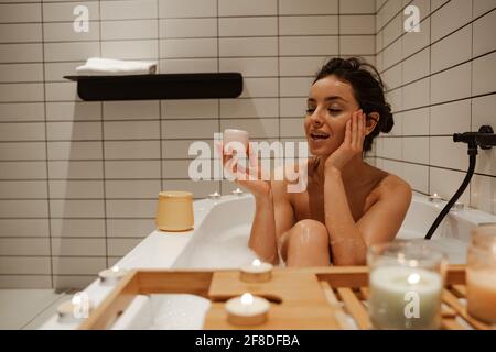 Young woman using cream on face, relaxing in the cosy bath full of foam and decorated with candles. Healthy treatment and enjoyment Stock Photo
