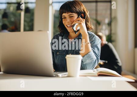 Cheerful businesswoman talking on cell phone while using laptop. Smiling young woman speaking on smart phone while looking at laptop in a coworking sp Stock Photo