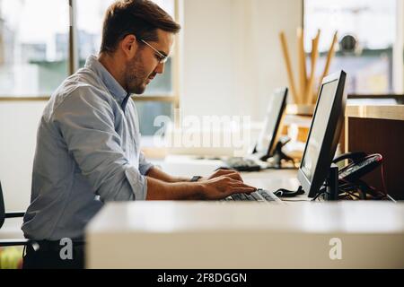 Freelancer typing on computer keyboard in office. Young man sitting at desk working on computer. Stock Photo