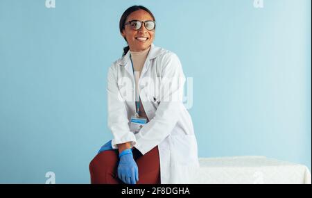 Portrait of a female doctor sitting on table and looking away. General practitioner wearing lab coat and hand gloves. Stock Photo